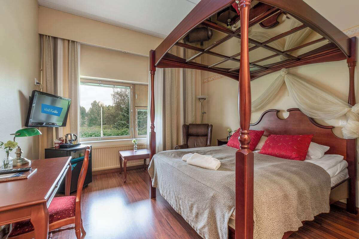 Una cama con dosel, un televisor en la pared, un escritorio con una silla y vistas al verde exterior.