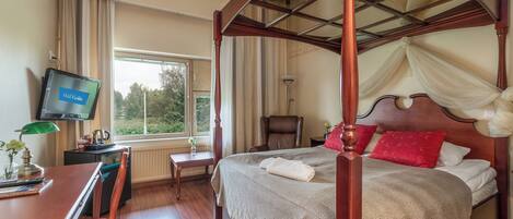 A four-poster bed with a canopy, a television on the wall, a desk with a chair, and a view of greenery outside.
