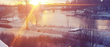 A winter landscape with a frozen river, bare trees, and a distant bridge during sunset.