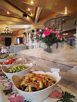 Uma área de buffet com diversos pratos, incluindo salada de massa e salada de frutas, em ambiente interno bem iluminado com lustre e decoração floral.