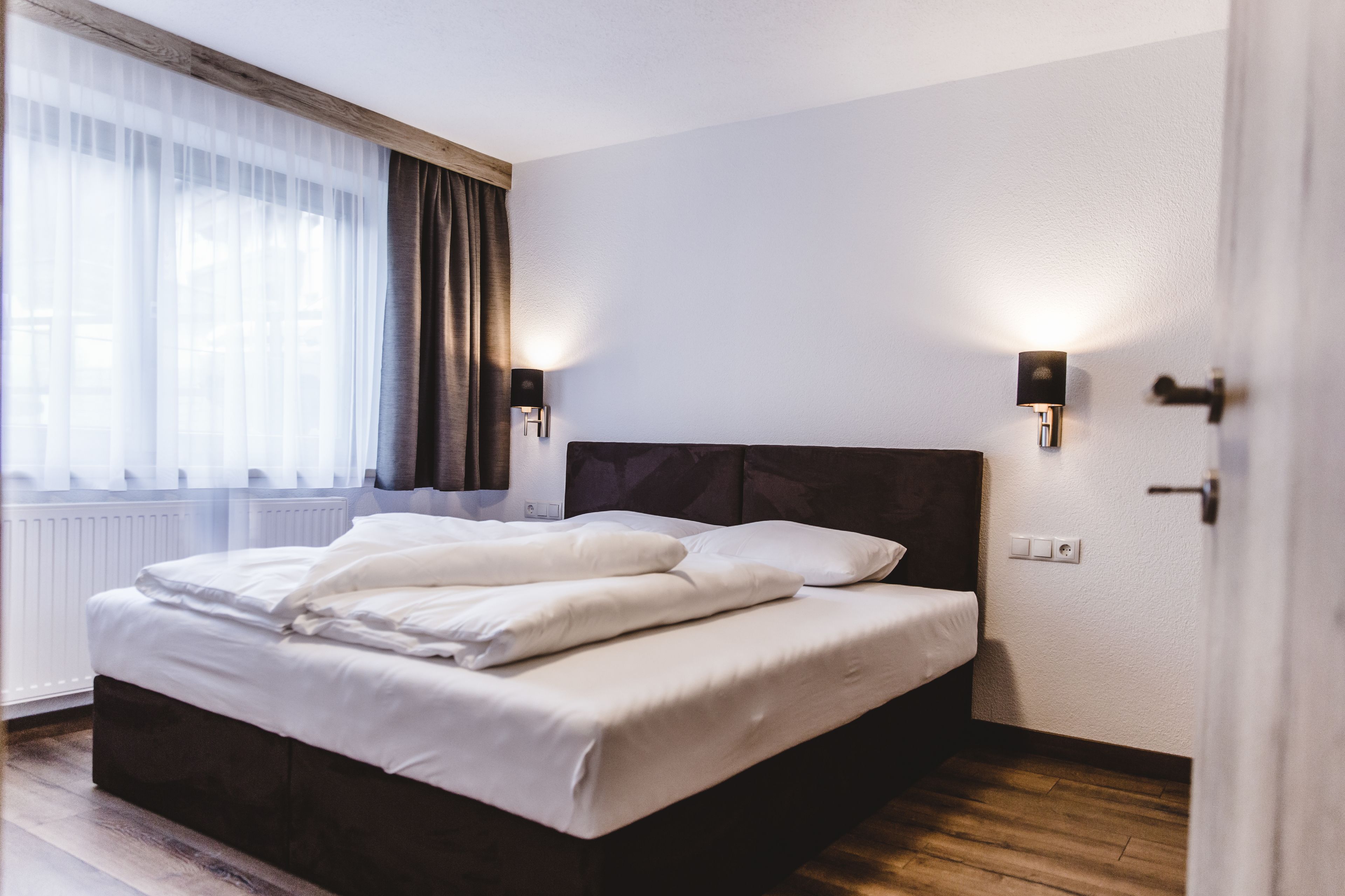 Een net opgemaakt bed met witte lakens, een donker hoofdbord en twee wandlampen.