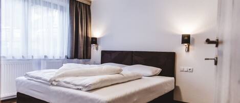 Een net opgemaakt bed met witte lakens, een donker hoofdbord en twee wandlampen.