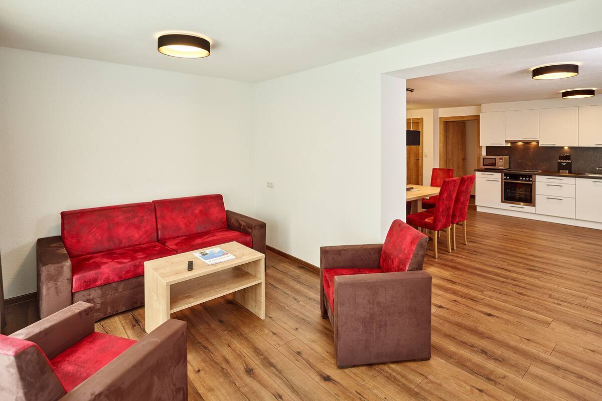 A modern living room with a red sofa, a wooden coffee table, and a red armchair. A kitchen is visible in the background.