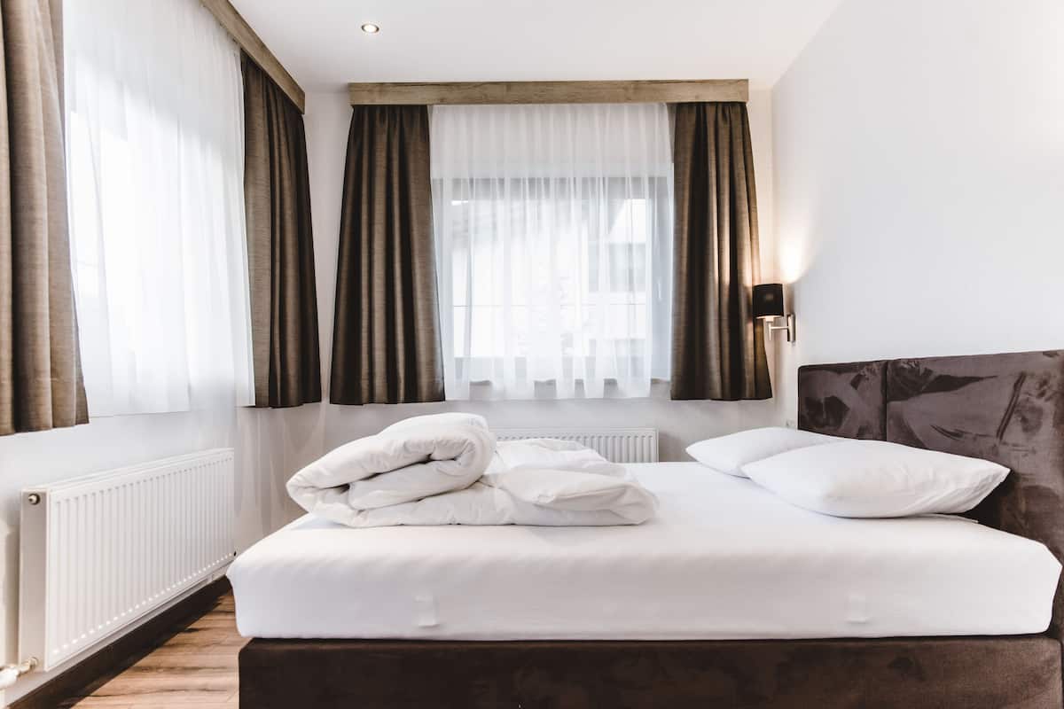 A hotel room with a bed, white linens, a radiator, and a window with curtains.