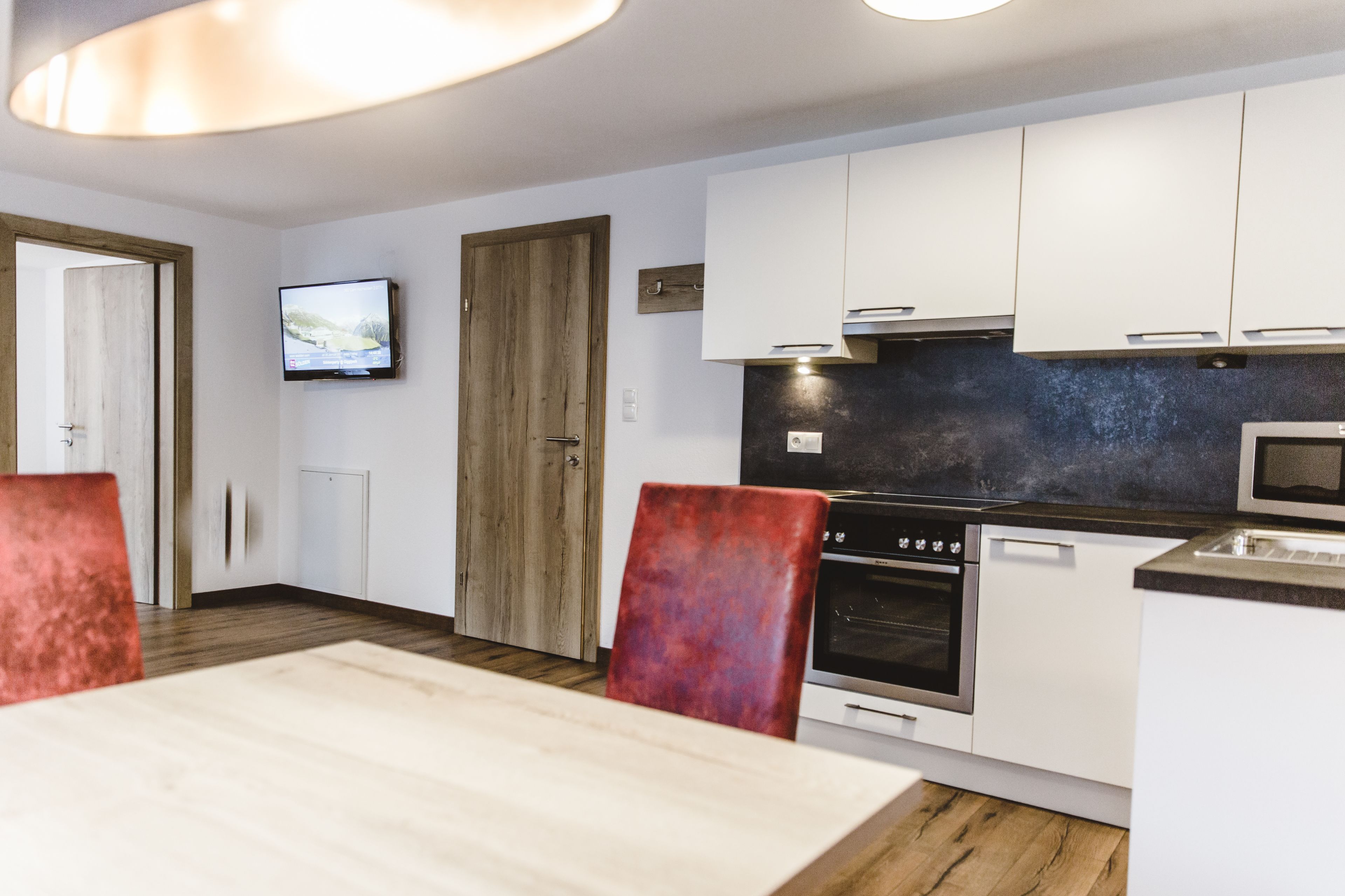 A modern kitchen with white cabinets, a wooden door, a TV mounted on the wall, and a dining area with a table and chairs.