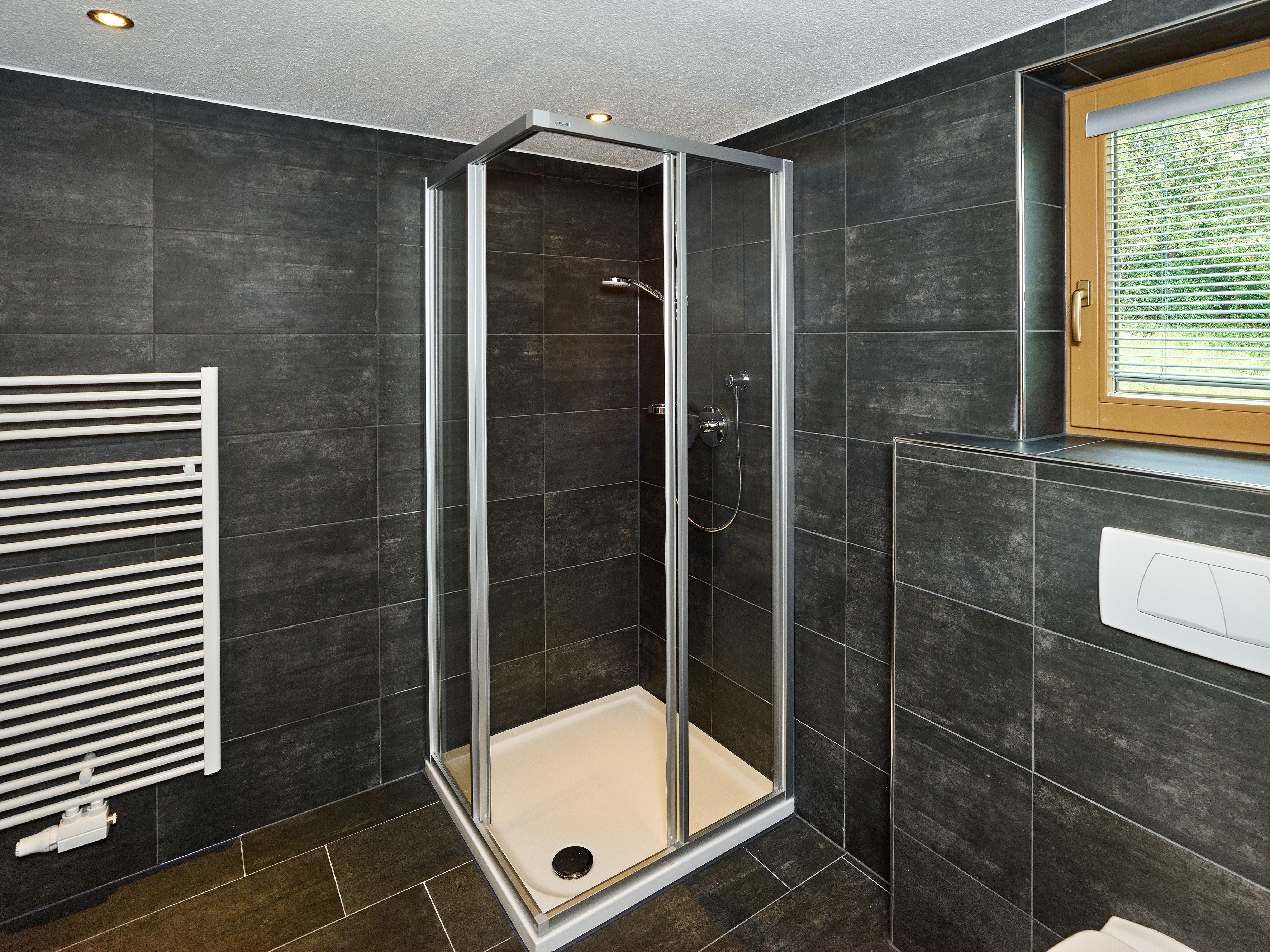 A modern bathroom with a glass-enclosed shower, dark tiled walls, a radiator, and a window with blinds.