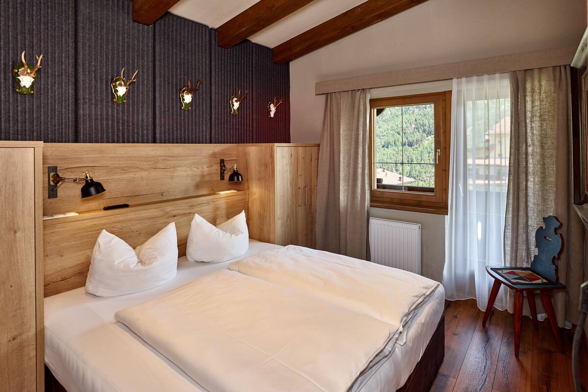 A bedroom with a wooden headboard, a large bed with white bedding, a nightstand, a lamp, a window with curtains, and a view of the outdoors.