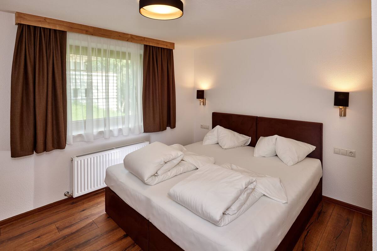 A neatly made bed with white linens and pillows, a wooden headboard, and two wall-mounted light fixtures.