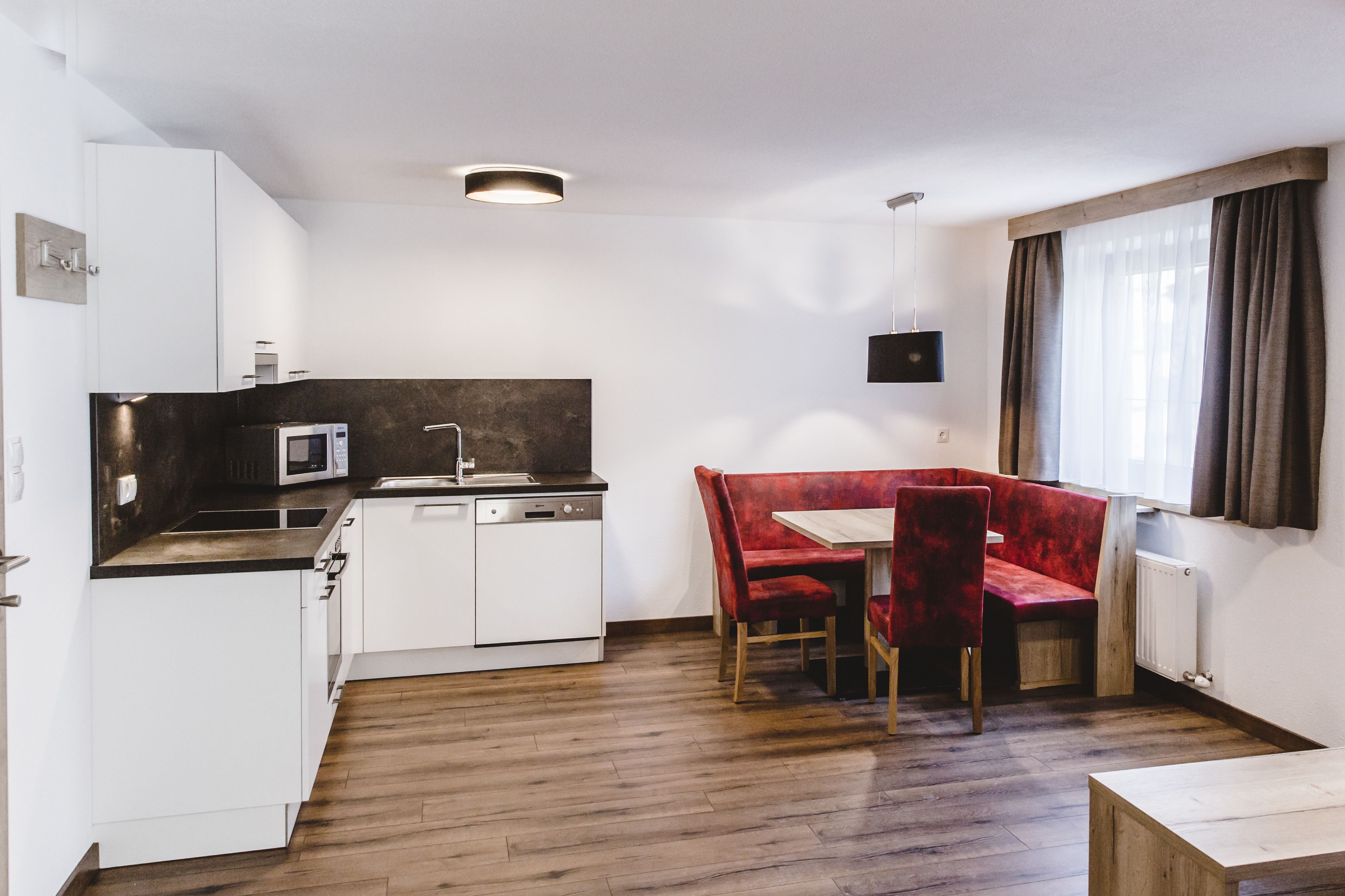 A compact kitchen with white cabinets, a microwave, and a dishwasher. There is a red upholstered bench with a table and chairs. A window with curtains is visible, and the room has wooden flooring.
