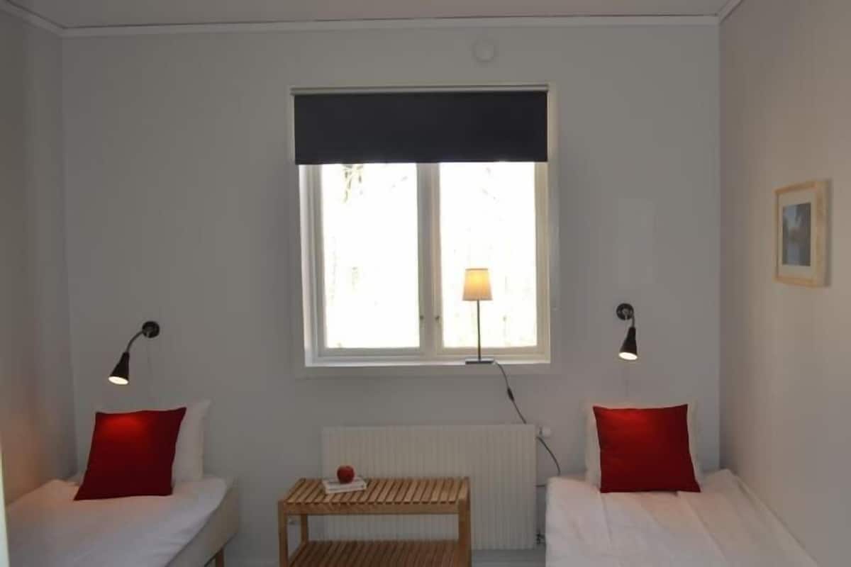 Una habitación pequeña y bien iluminada con dos camas individuales, una mesita y dos lámparas de noche.