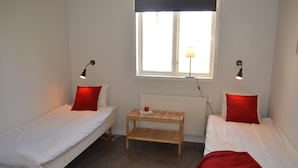 Une petite chambre propre, avec deux lits simples, une table de chevet en bois, une fenêtre donnant sur un beau paysage et deux lampes fixées au mur.