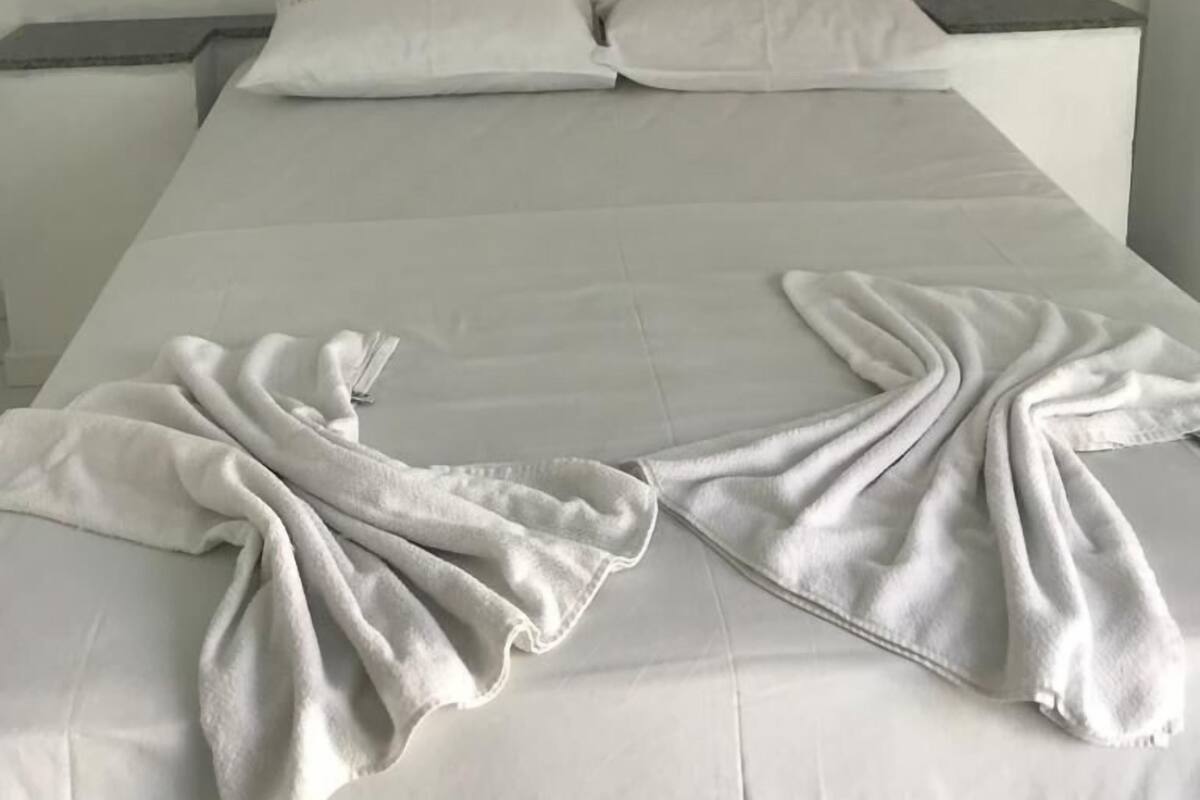 A bed with white linens and a blanket, a painting of a ship on the wall, and a bedside table.
