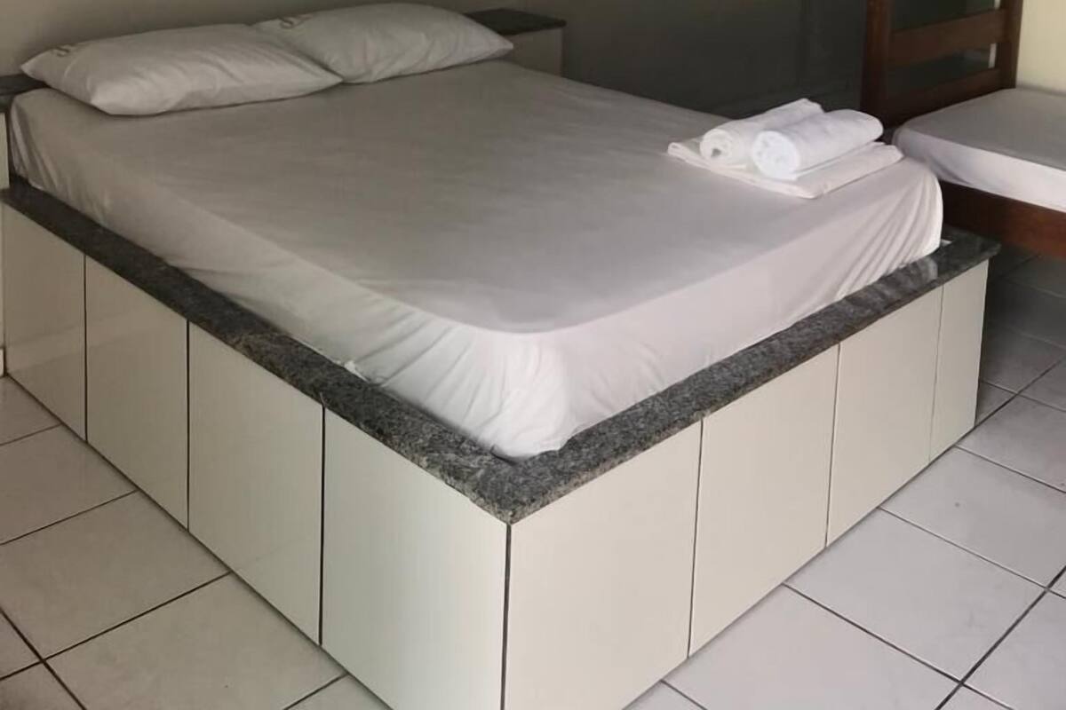 A hotel room with a bed, a painting of flowers, an air conditioning unit, and a wooden bunk bed.
