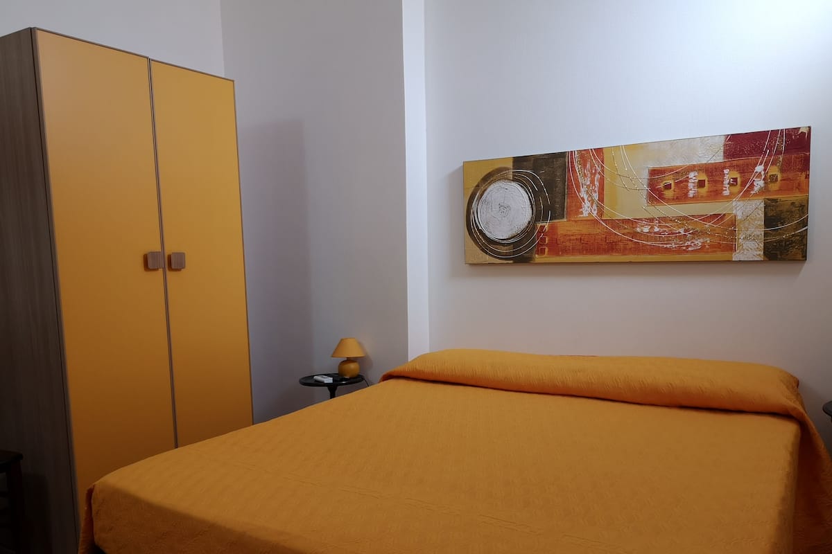 A room with a bed, a wardrobe, a small table with a lamp, and a painting on the wall.