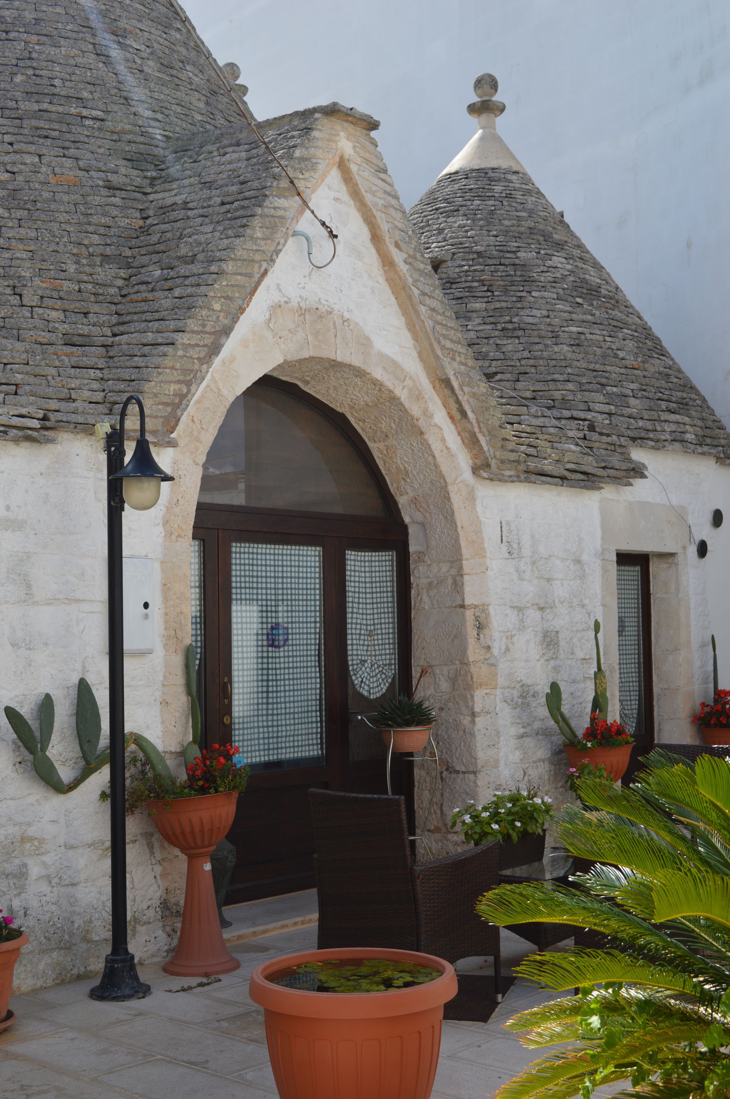 A building with a stone facade, a wooden door, and a tiled roof. There are potted plants and a street lamp.
