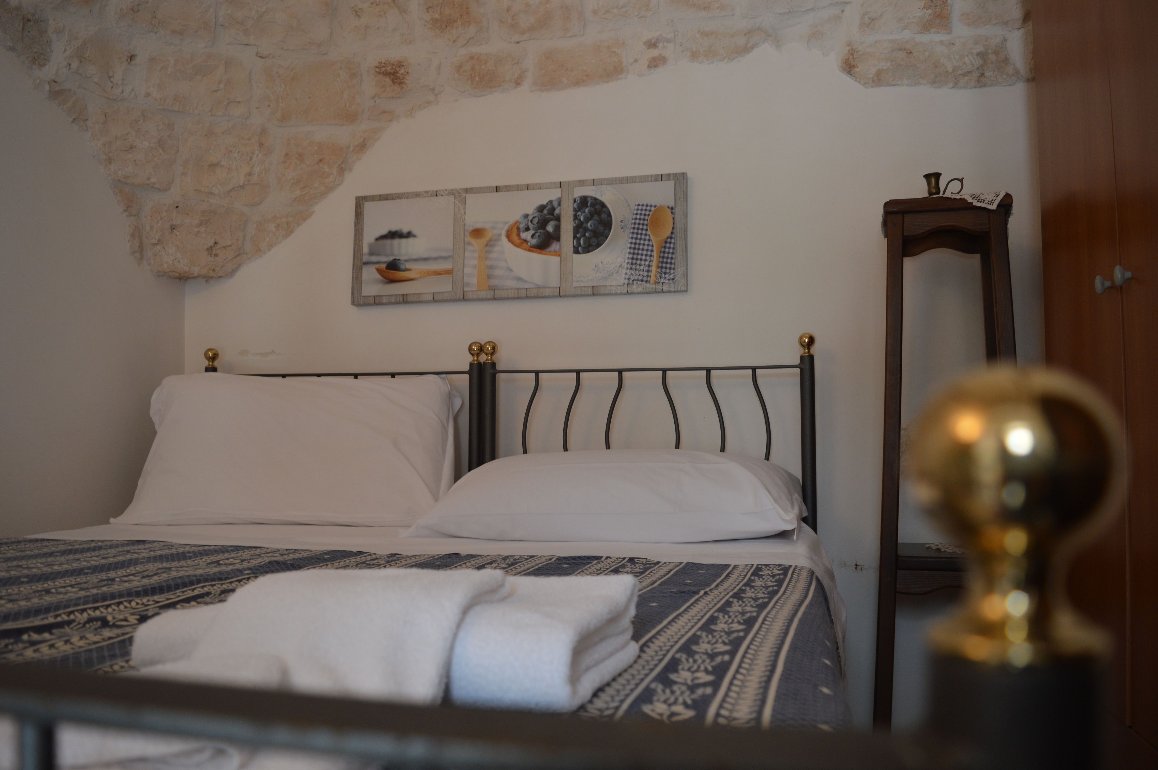 A bed with white linens and a patterned blanket, a wall-mounted picture of kitchen utensils, and a wooden nightstand with a lamp.