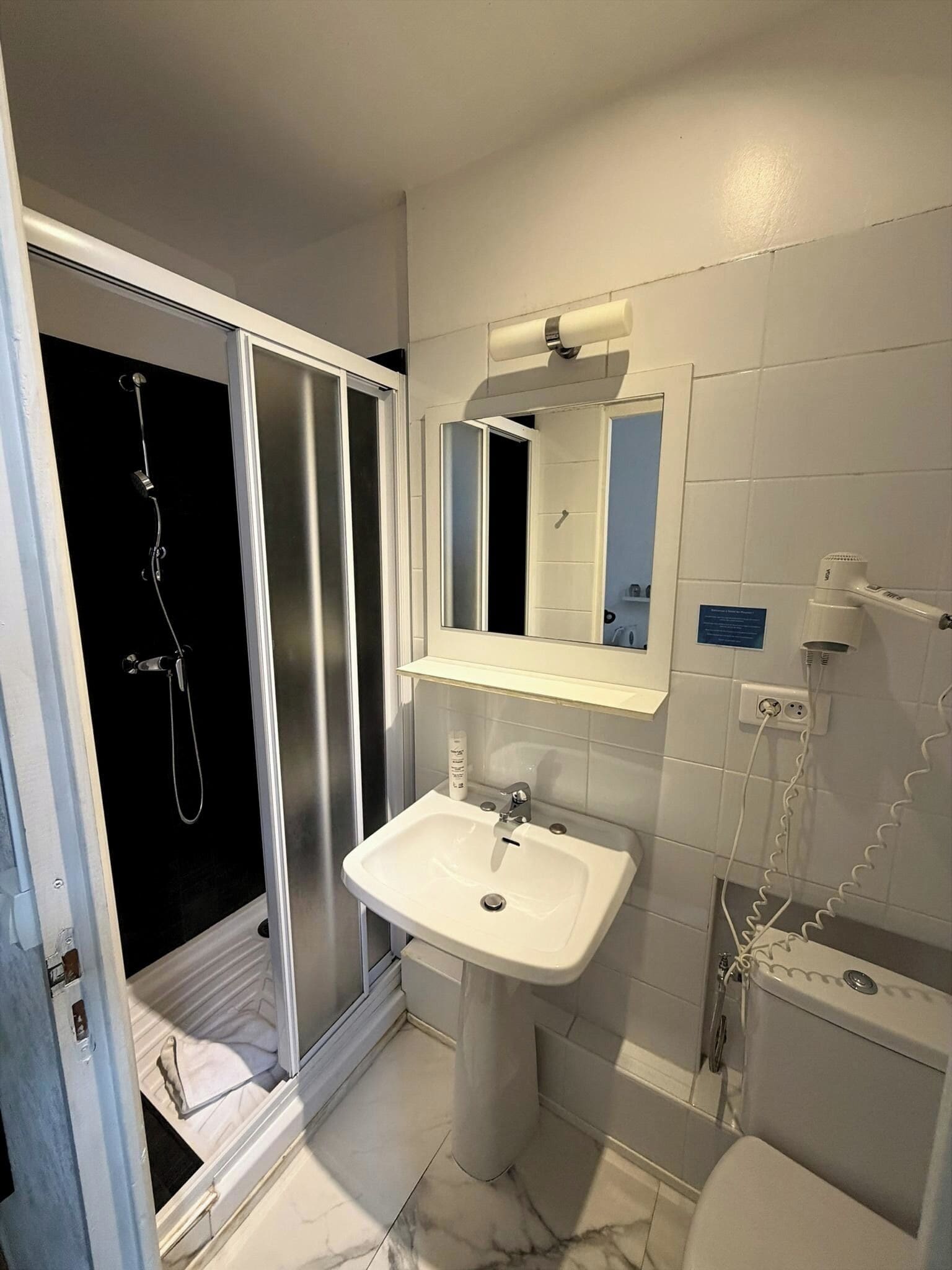 Un baño moderno con cabina de ducha de vidrio, lavamanos pedestal, espejo y secador de pelo en la pared.