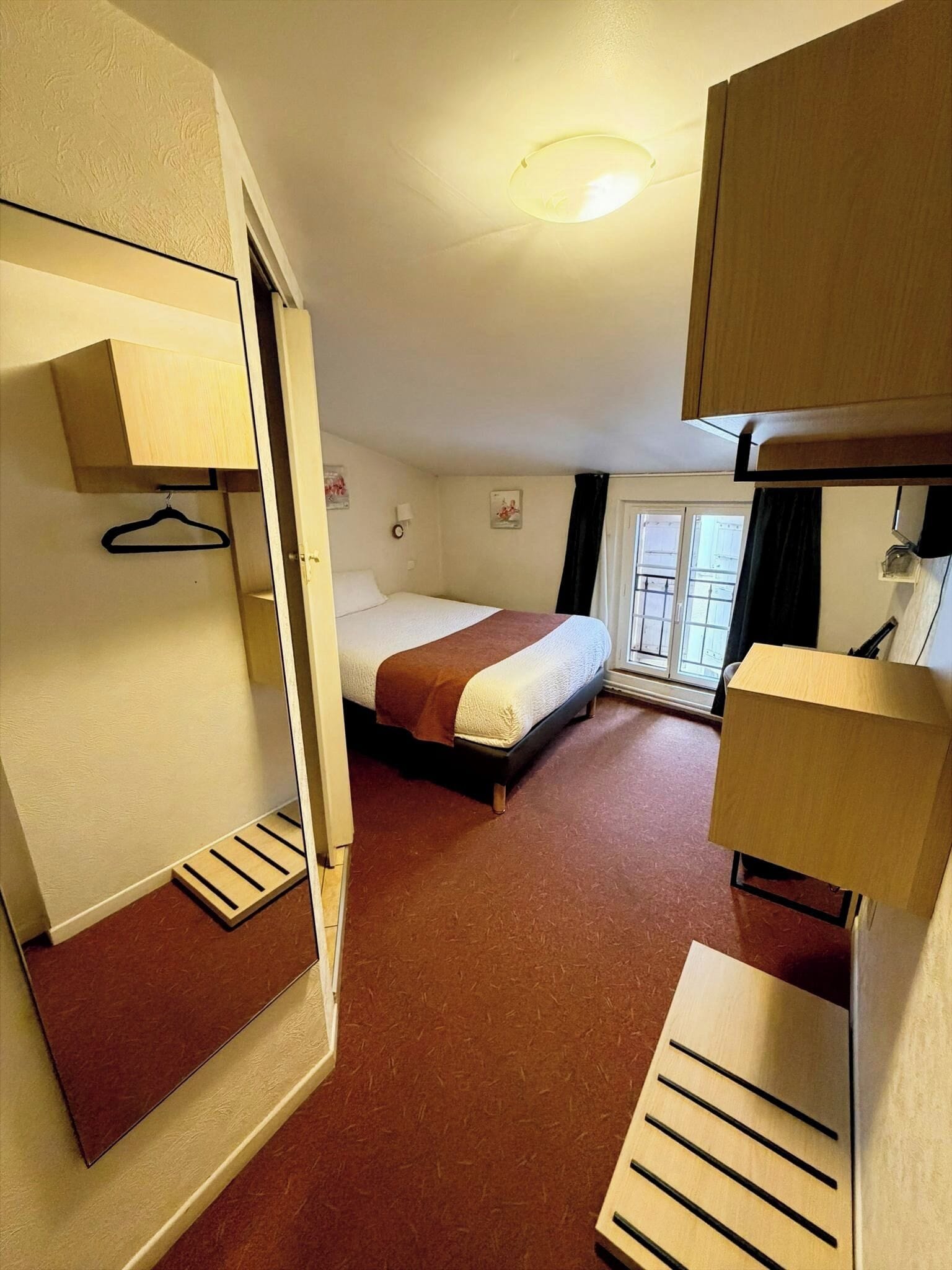 Une chambre d’hôtel de taille modeste, comprenant un lit simple, un petit bureau, un porte-manteau et une fenêtre avec des rideaux.