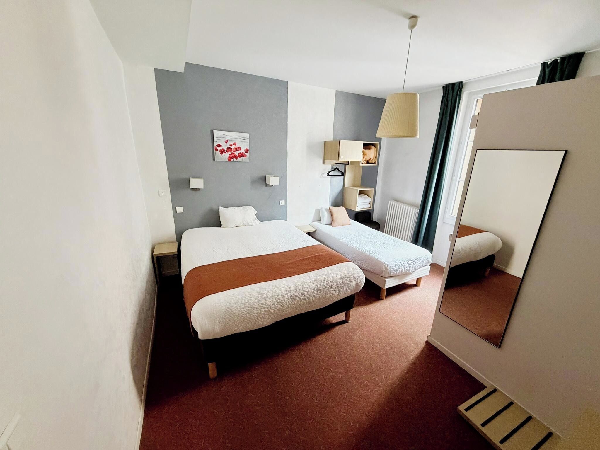 A hotel room with two beds, a large mirror, a bedside table, a lamp, and a painting on the wall.