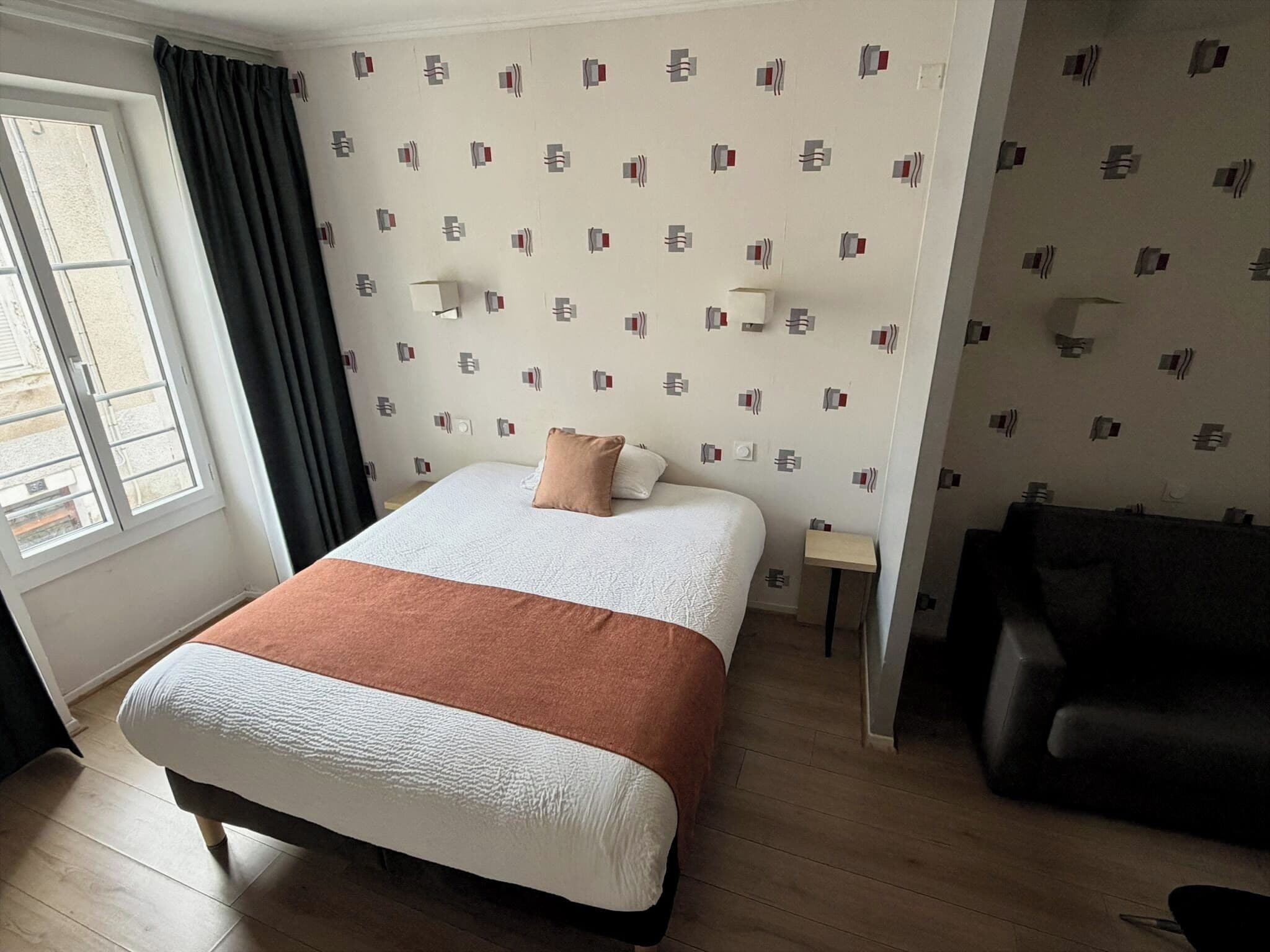A hotel room with a bed, a small bedside table, a lamp, a sofa, and a window with curtains.