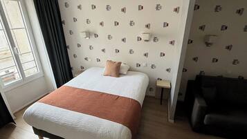 A hotel room with a bed, a small bedside table, a lamp, a sofa, and a window with curtains.
