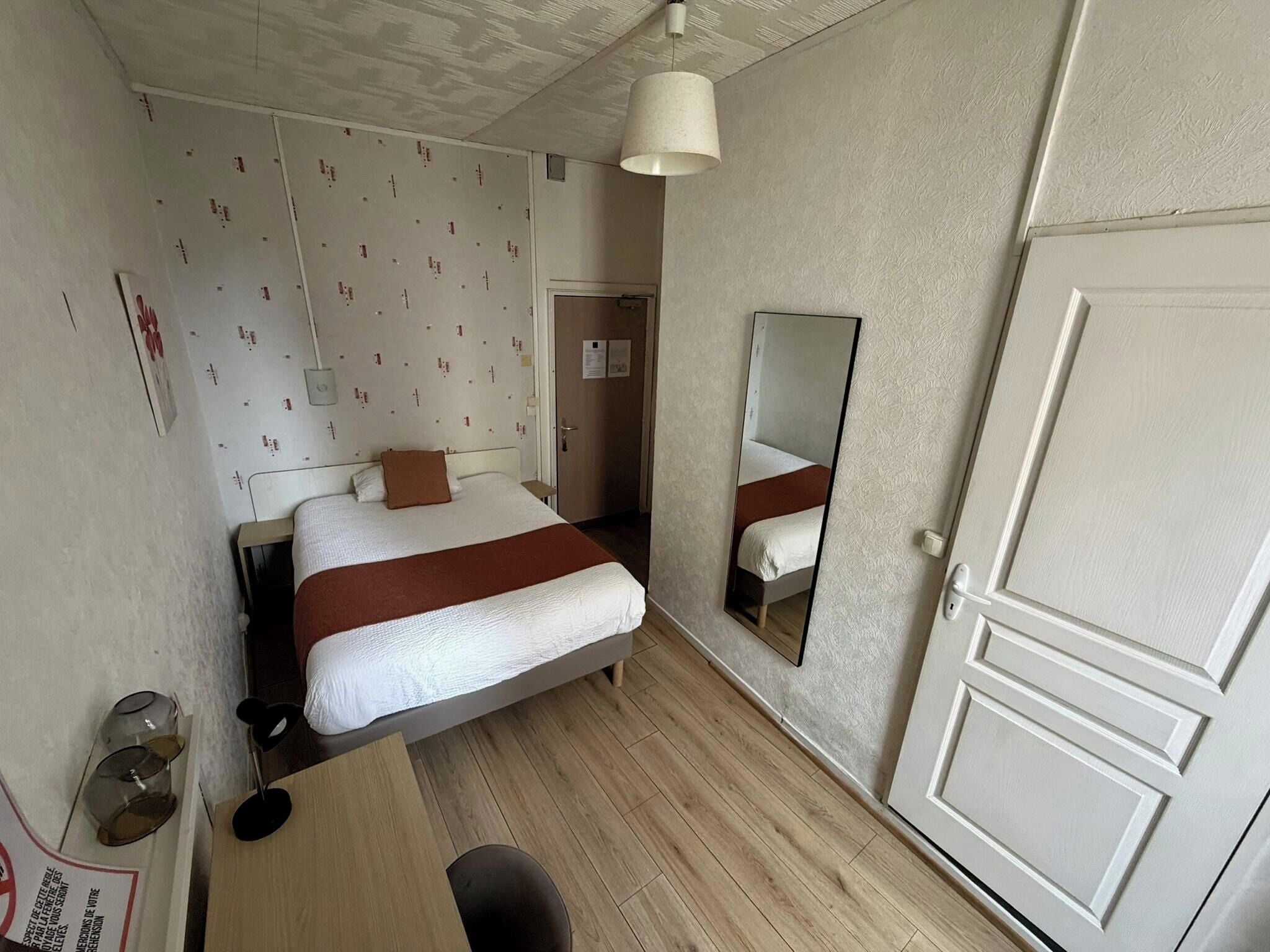 A hotel room with a bed, a desk, a chair, a mirror, and a door.