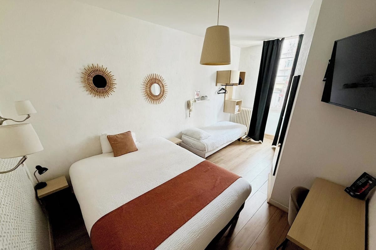 Ein Hotelzimmer mit zwei Betten, einem Schreibtisch, einem Fernseher und einem Fenster mit Vorhängen.