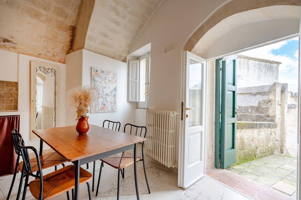 A dining area with a wooden table, chairs, and a vase of dried flowers. There is a radiator, a door leading outside, and a window.