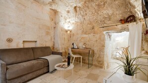 A cozy living room with a sofa, a small table with a bottle and glasses, a desk with a chair, and a potted plant. - La Corte Vetere (Matera)