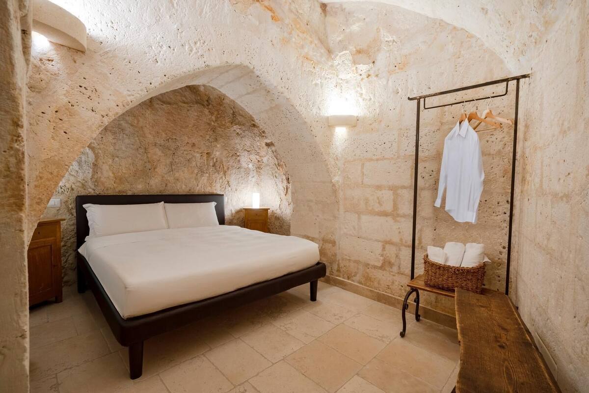 A bedroom with a stone arched ceiling, a bed with white linens, a wooden bench, and a clothing rack with hangers.