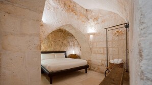 A bedroom with a stone wall, a bed, a nightstand, a wooden bench, and a hanging light fixture. - La Corte Vetere (Matera)