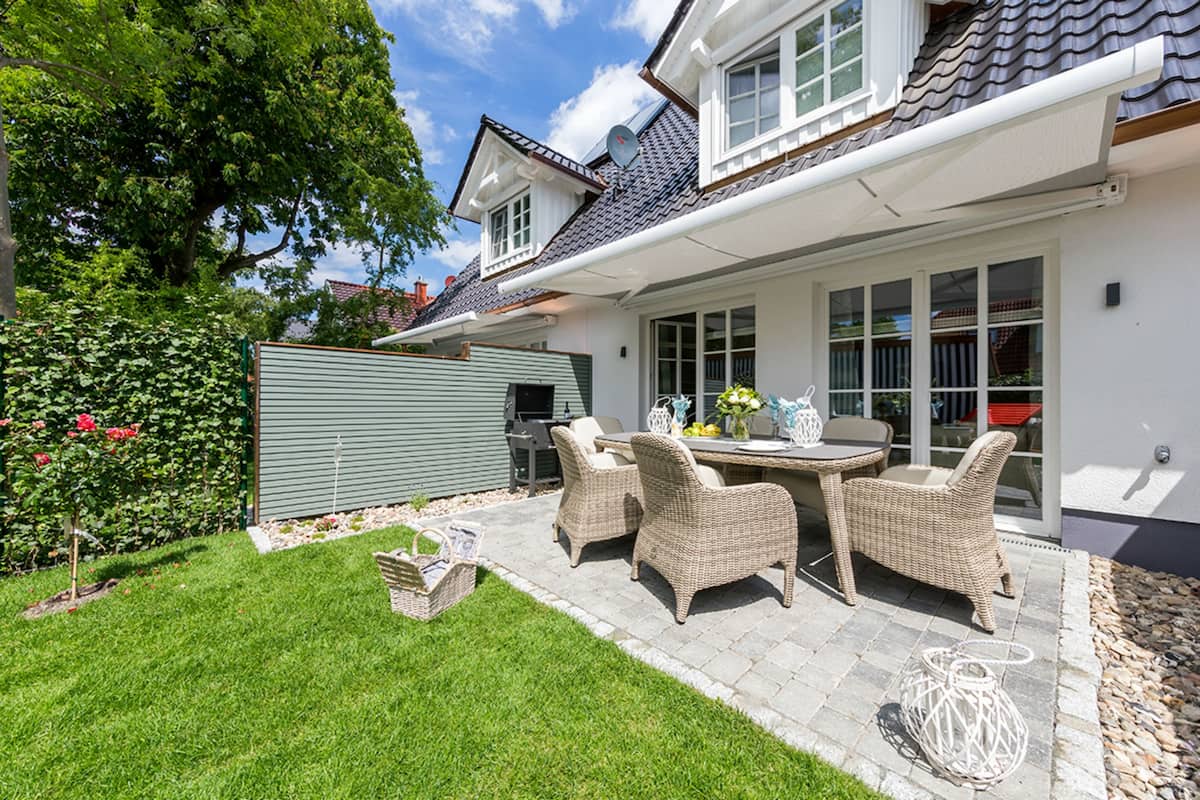 Een terras met rieten meubilair, een barbecue en een tuin met een haag en bloemen.