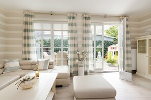 A modern living room with a sofa, a coffee table, and a dining area. There is a large window with curtains, a view of the outside, and a cabinet. - Ferienhäuser Zingster Perlen & TraumZeit (Zingst)