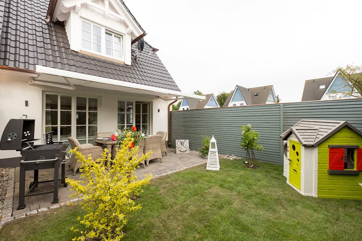 Een achtertuin met een barbecue, rieten meubilair, een kleine schuur en een groene speeltuintje.