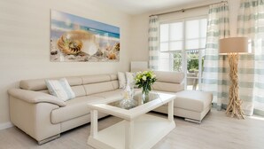 Uma sala de estar com um sofá bege, uma mesa de centro de vidro e um quadro de uma concha na parede.