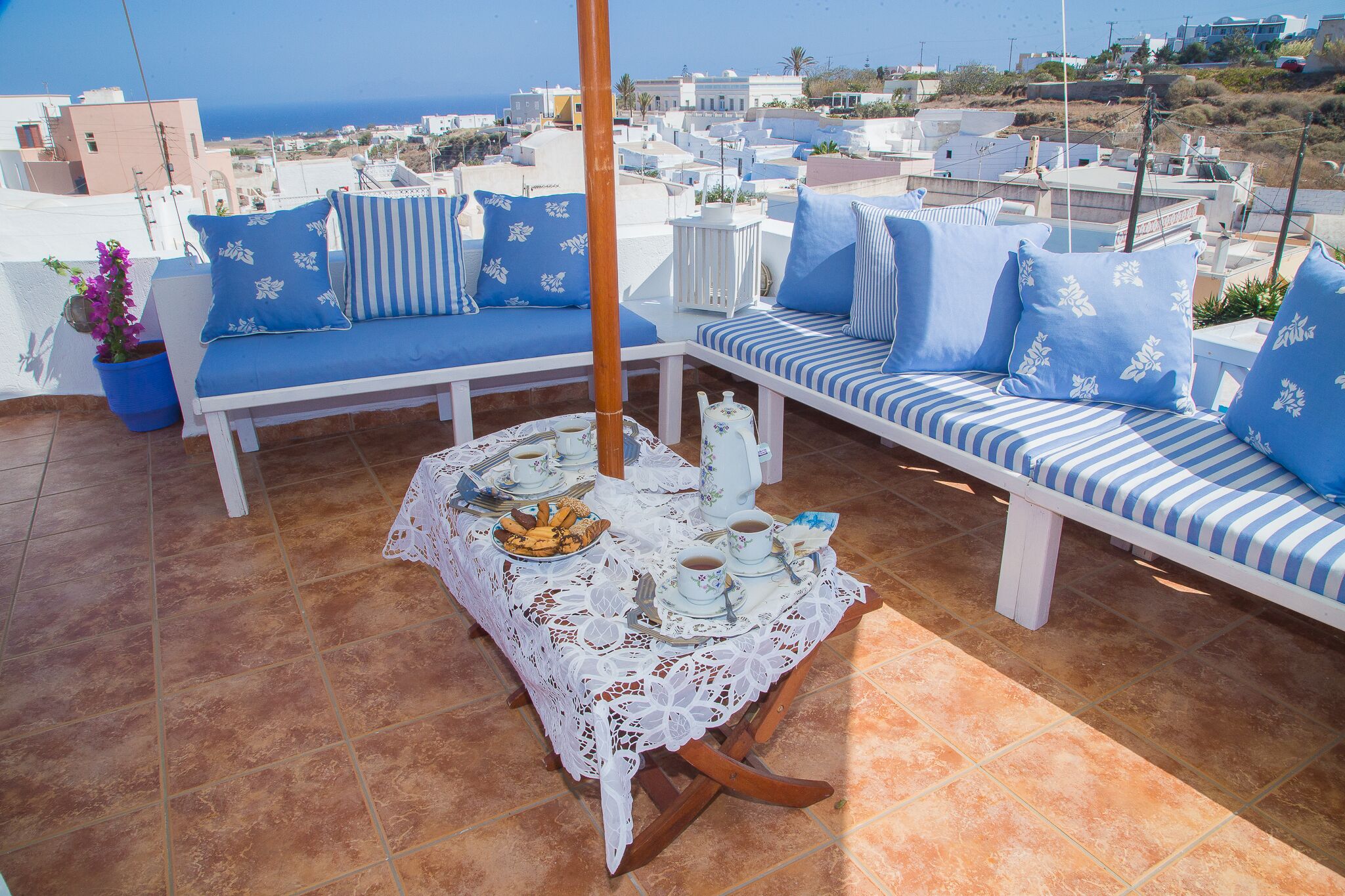 A terrace with a white and blue striped sofa set, a table with a lace tablecloth, and a view of the sea and buildings.