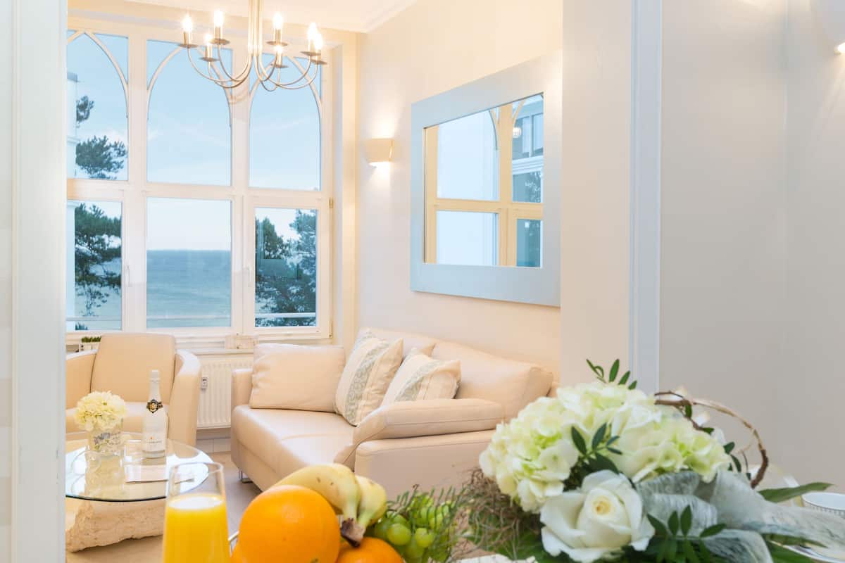 Una habitación con vistas al mar, un sofá, una mesa de vidrio con una botella y vasos, y un ramo de flores.