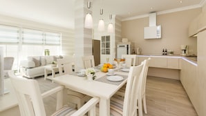 A modern, well-lit dining area with a white table set for four, surrounded by white chairs. A kitchen area is visible in the background with a white countertop and modern appliances.