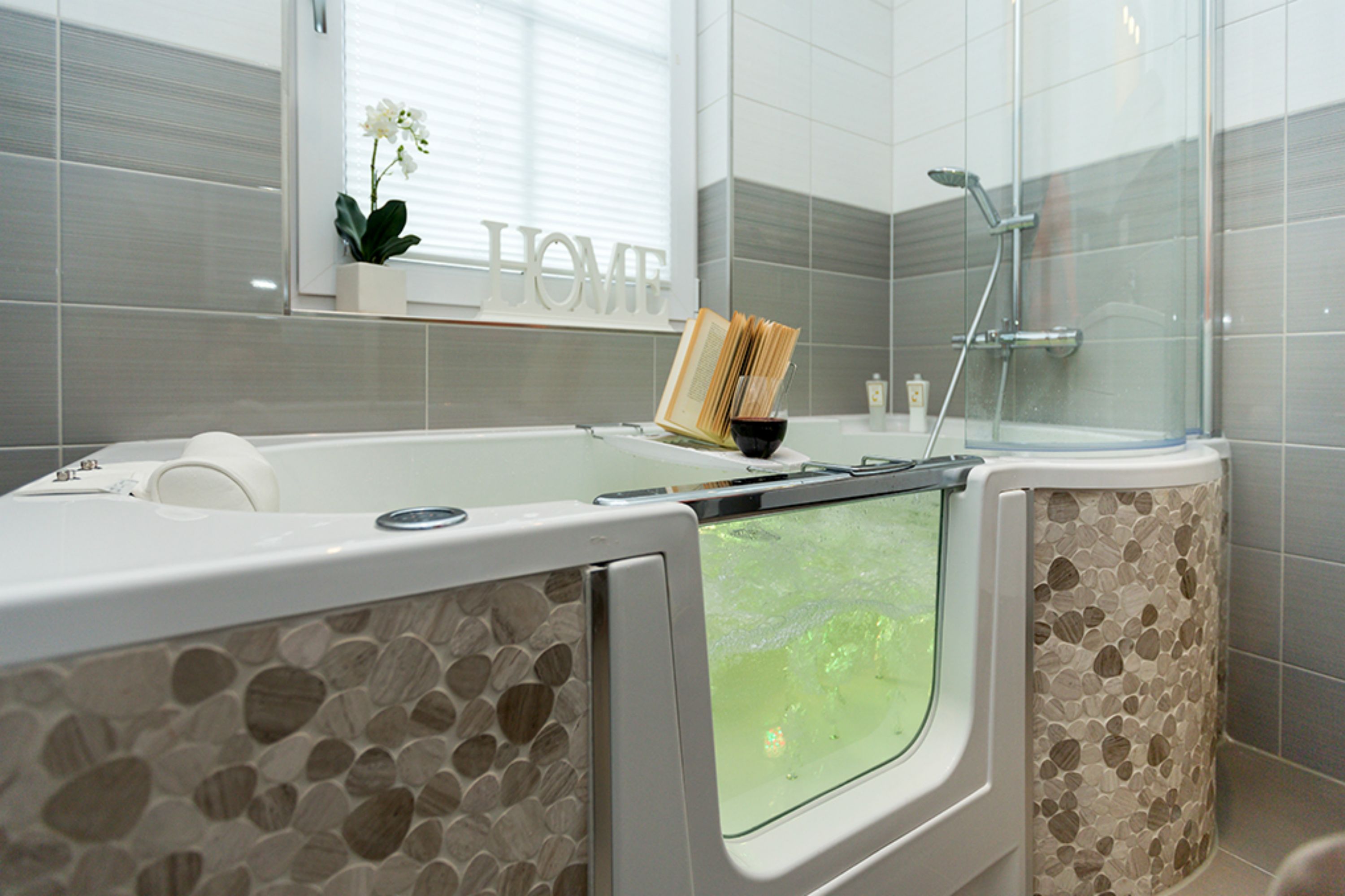 A modern bathroom with a stone-tiled shower enclosure, a bathtub with a glass window, and a small potted plant on the windowsill.