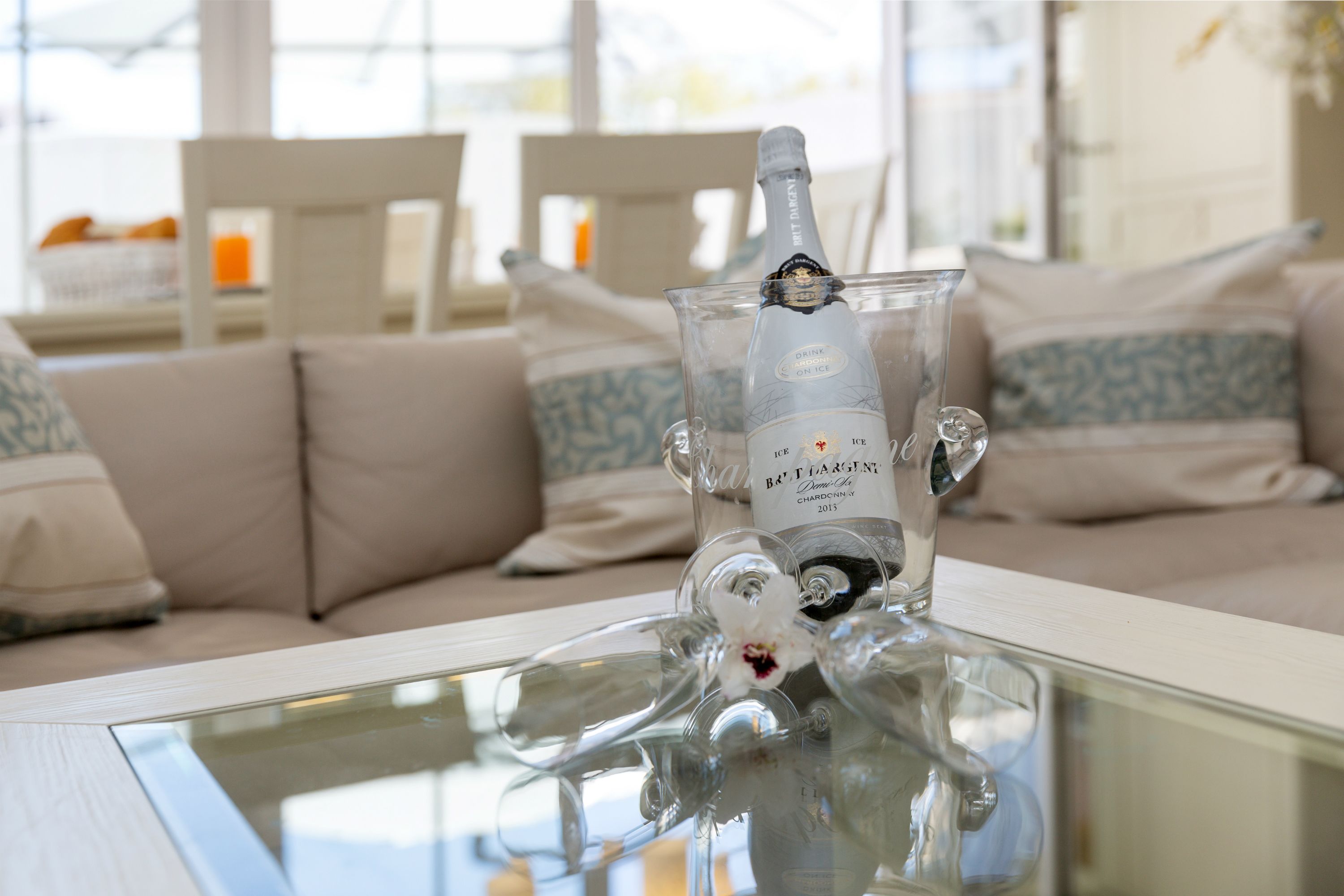 A bottle of wine on a glass table with a sofa and cushions in the background.