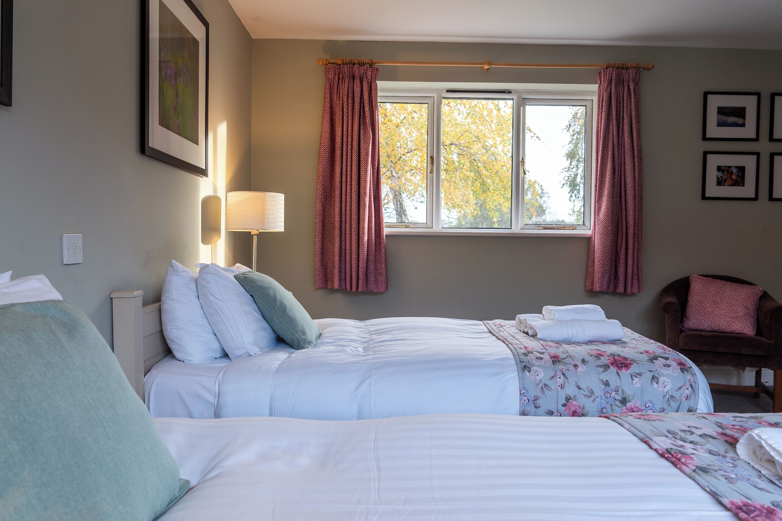 Quarto de hotel com duas camas, uma janela com cortinas, uma cadeira e quadros na parede.