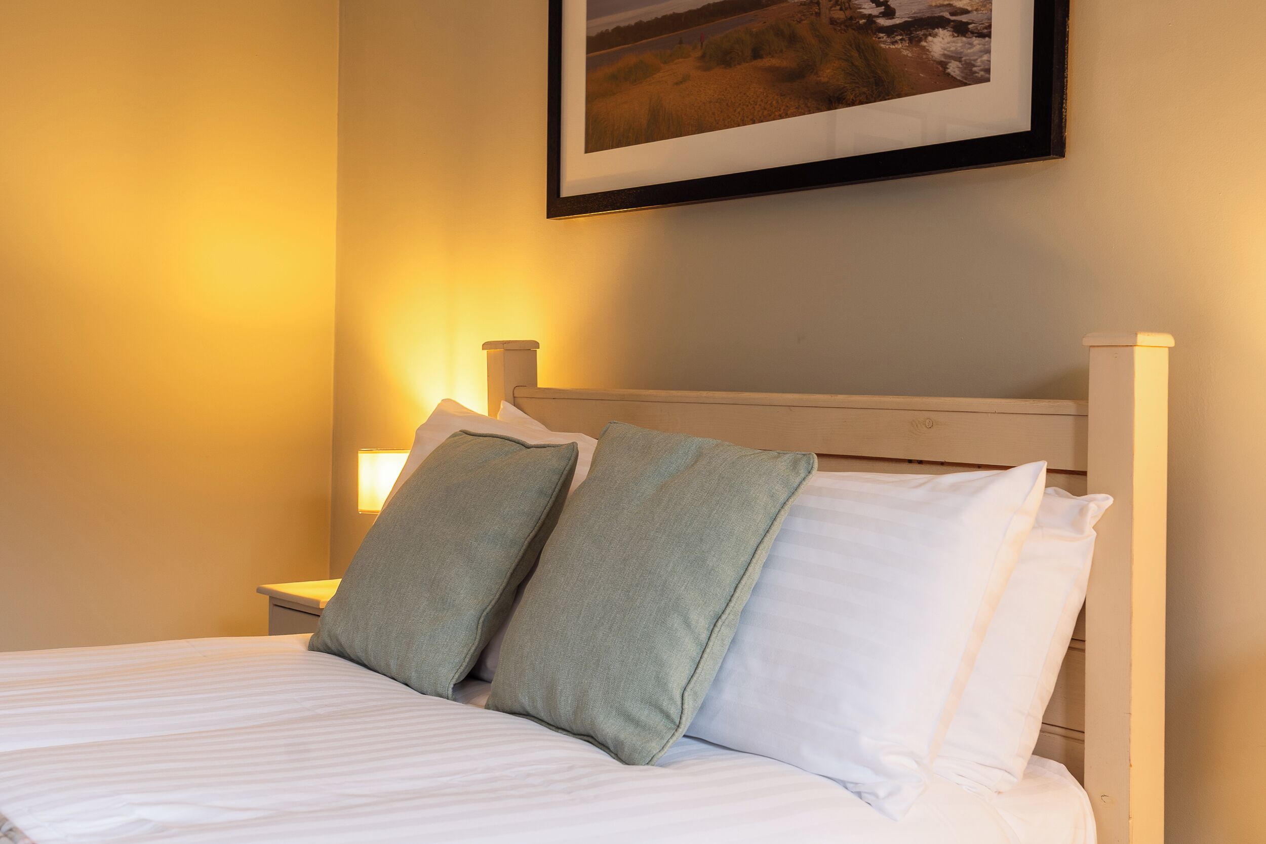 A neatly made bed with two pillows, a framed picture on the wall, and a bedside lamp.