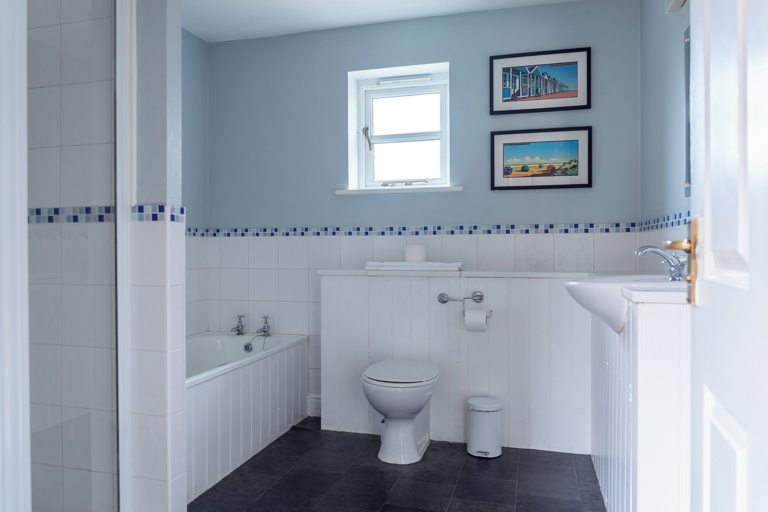 A bathroom with a toilet, sink, and bathtub. There is a window above the toilet and two framed pictures on the wall.