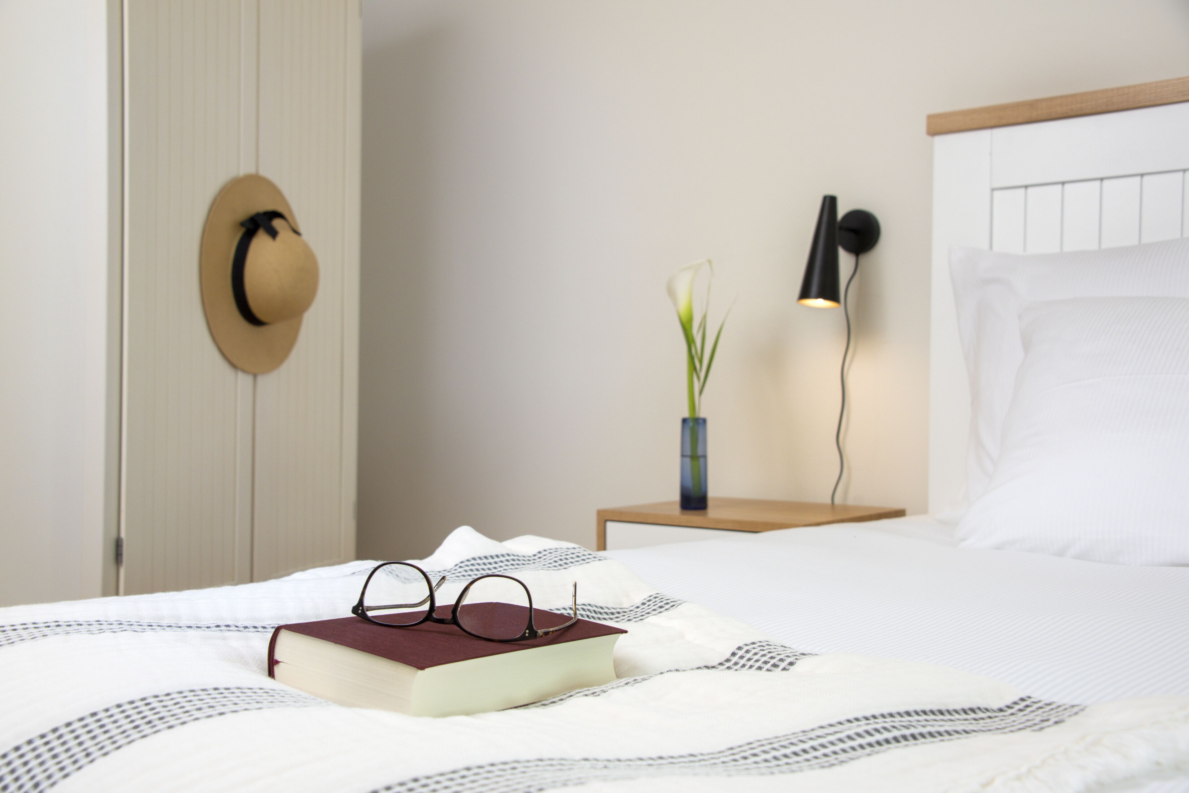 A neatly made bed with a book, glasses, and a hat on the side. There is a nightstand with a lamp and a vase with a plant.
