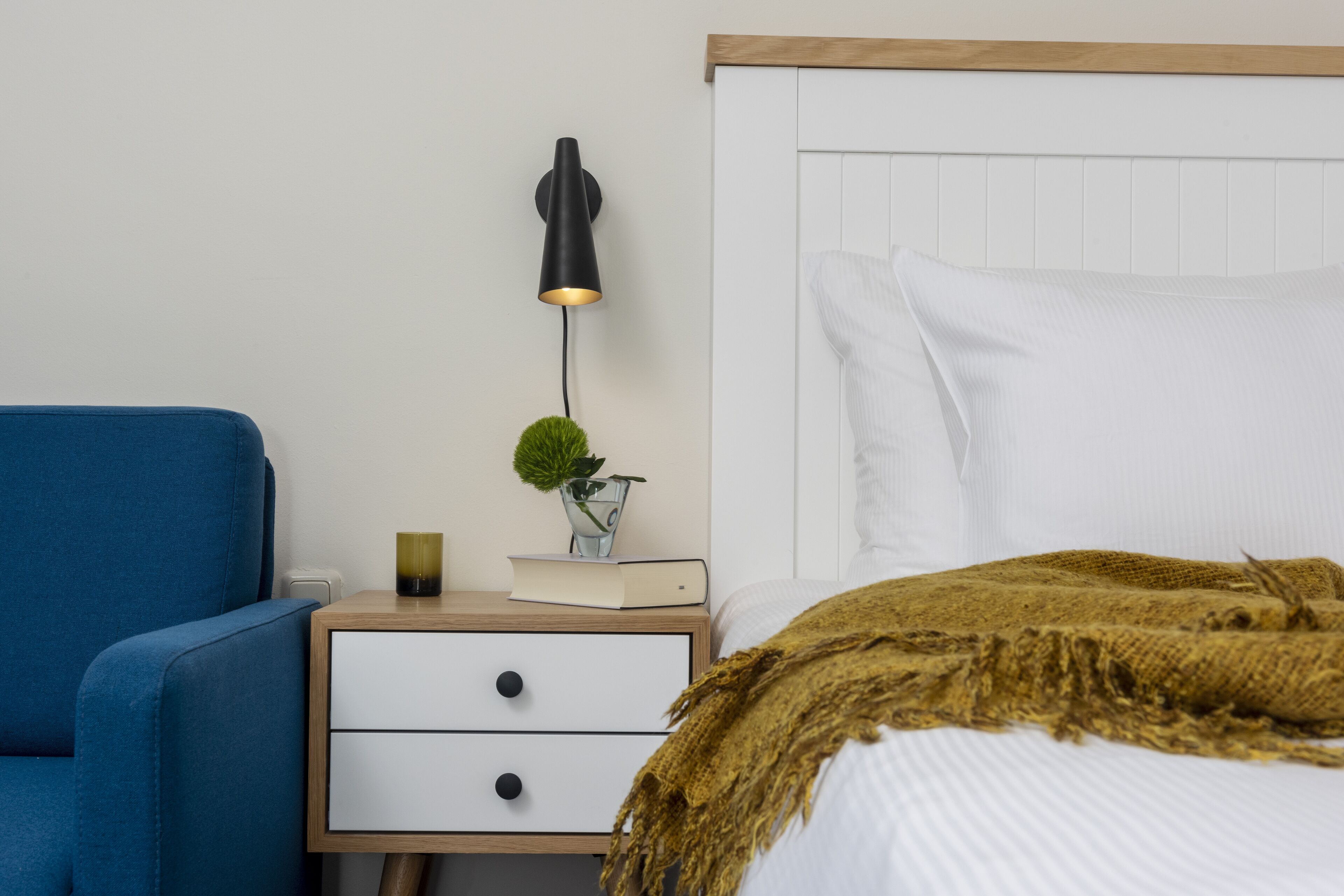 Un dormitorio con una cama, una mesita de noche con lámpara, una silla azul y una planta pequeña.