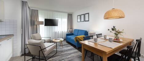 Una sala de estar moderna con un sofá azul, una mesa de comedor con sillas, un televisor montado en la pared y una zona de cocina con estufa.