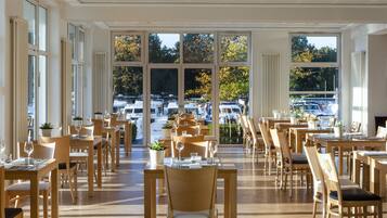 A spacious dining area with large windows, wooden tables and chairs, and potted plants.