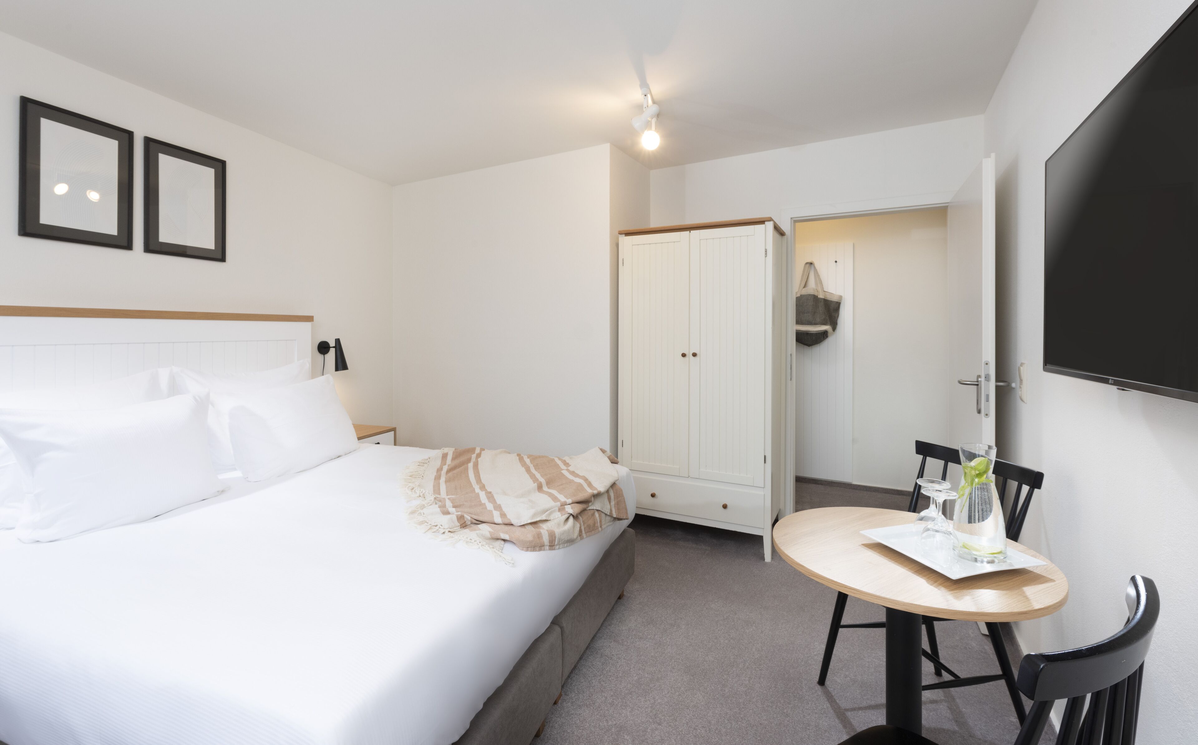 Een hotelkamer met een bed, nachtkastje, stoel, rond tafel, kledingkast en een aan de muur bevestigde televisie.