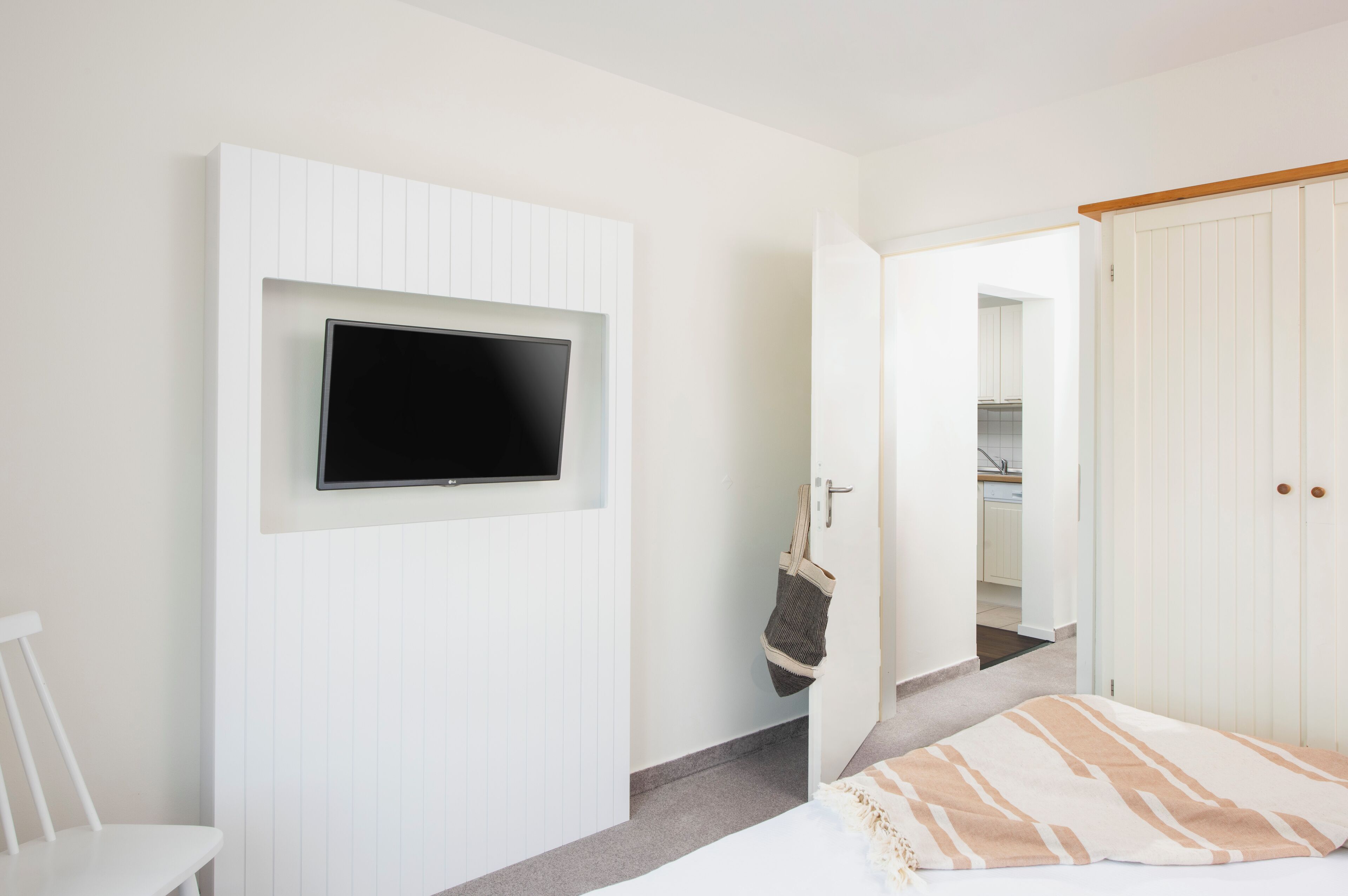 A bedroom with a bed, a TV mounted on the wall, a door leading to a bathroom, and a white wardrobe.