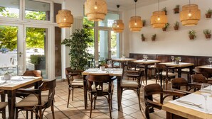 Un interior de restaurante con varias mesas, lámparas colgantes de mimbre, plantas en macetas y amplios ventanales que dejan entrar luz natural.